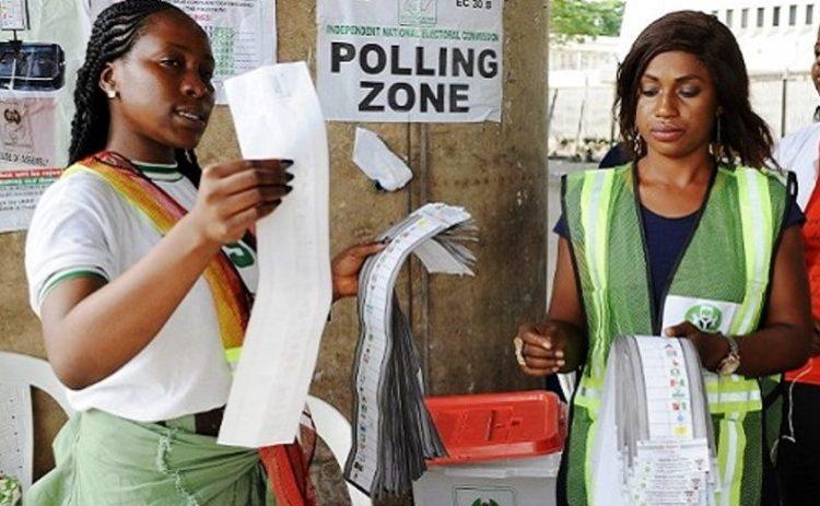Two female NYSC corps members serving as INEC ad-hoc polling staff sorting ballot papers at an official polling zone in Nigeria.