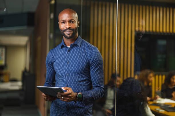 A confident male cybersecurity specialist holding a digital tablet in a corporate tech office setting.