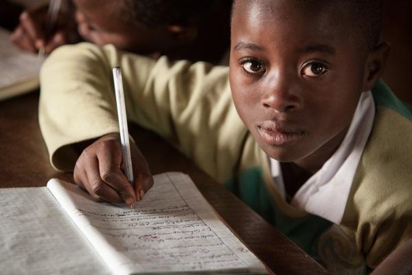 A young Nigerian primary school student focused on writing in his notebook during a classroom lesson.