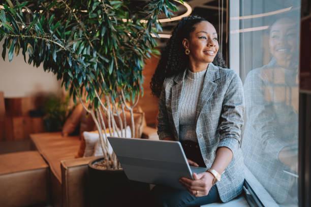 A smiling professional Black woman holding a laptop while sitting by a large office window.
