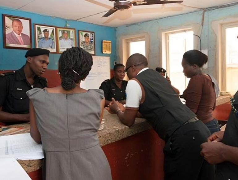 Civilians standing at a charge counter inside a Nigerian police station interacting with officers under official portraits.