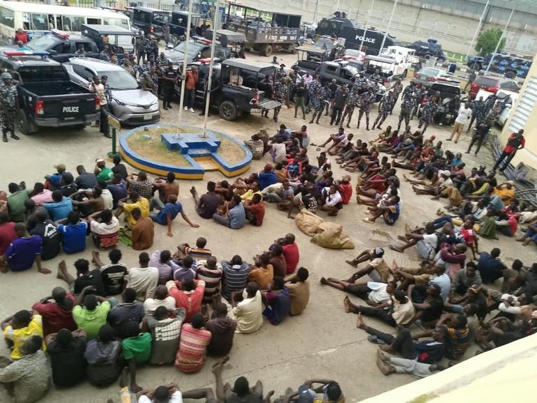 Detained individuals sit together on the ground outside a police station.