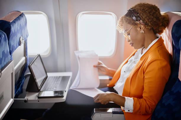 A Black businesswoman in a bright orange blazer working on documents while traveling in the premium cabin seat of a private jet.