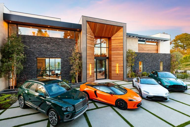 A modern luxury mega-mansion driveway packed with multiple exotic supercars, including a Bentley SUV, a McLaren, and an Aston Martin at sunset.
