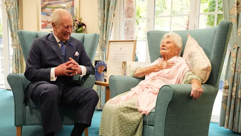 King Charles III sitting and smiling with 117-year-old Ethel Caterham, the world's oldest person, in a bright care home room.