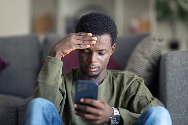 Worried young African man holding his hand to his forehead while checking his phone messages on a couch.