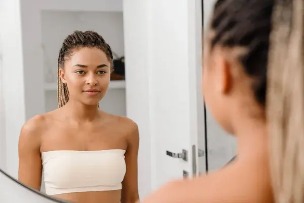 A young Black woman standing and looking thoughtfully at her reflection in a mirror.