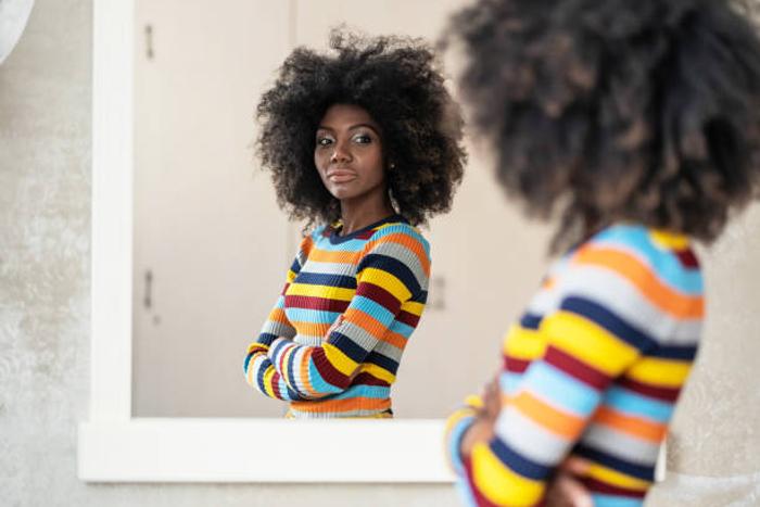 A young Black woman with an afro hairstyle standing with folded arms and looking thoughtfully at her reflection in a mirror.