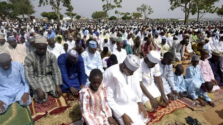 A large outdoor congregation of Nigerian Muslim men and children dressed in traditional attire performing Eid prayers on prayer mats.