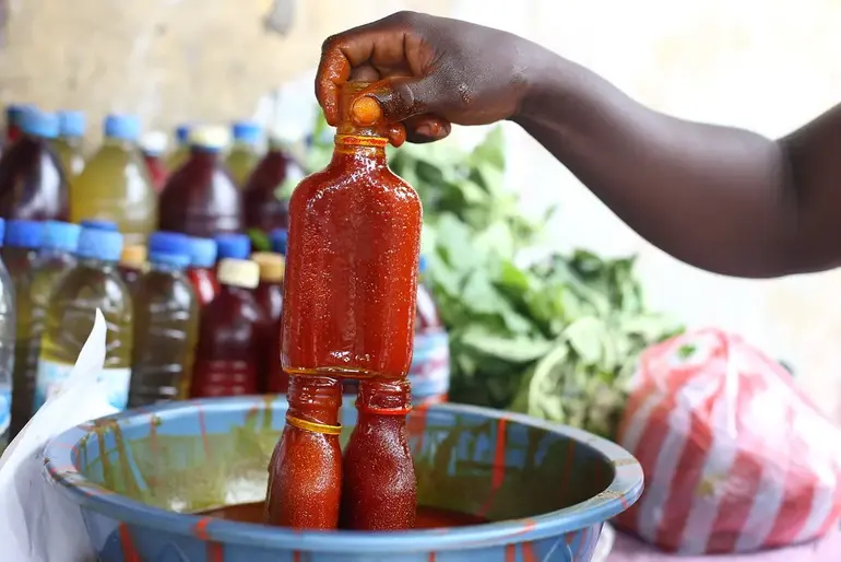 A vendor in a Nigerian market lifting a clear plastic bottle filled with thick, deep red palm oil from a large basin.