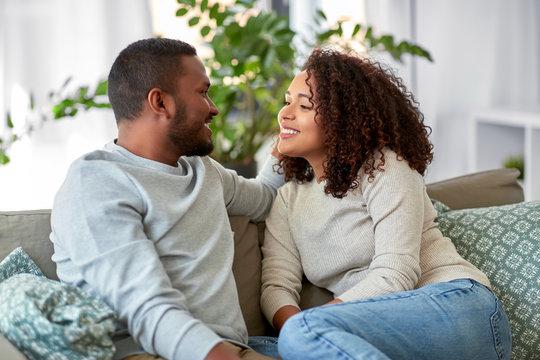 A young Black man and woman sitting close together on a living room couch, sharing a warm, loving glance.