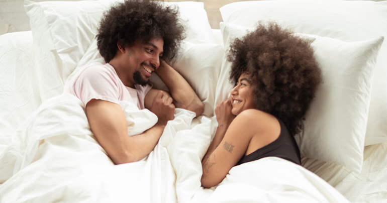 A happy Black couple lying in a cozy white bed, smiling and looking into each other's eyes while talking.