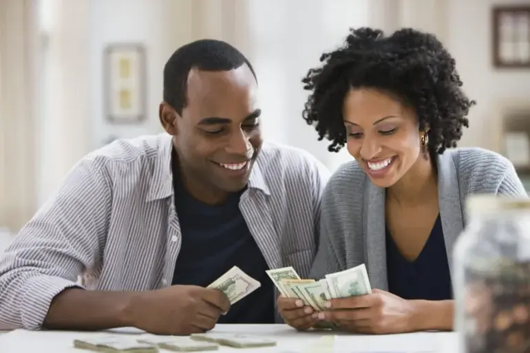 A smiling Black couple sitting at a table together counting US dollar bills.