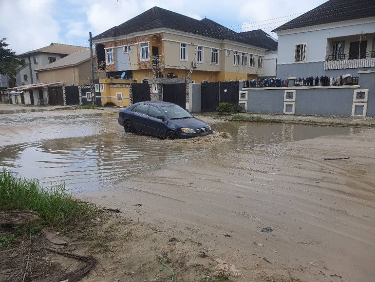 A dark sedan car attempting to drive through a large, muddy pool of floodwater in a residential estate in Lekki, Lagos.