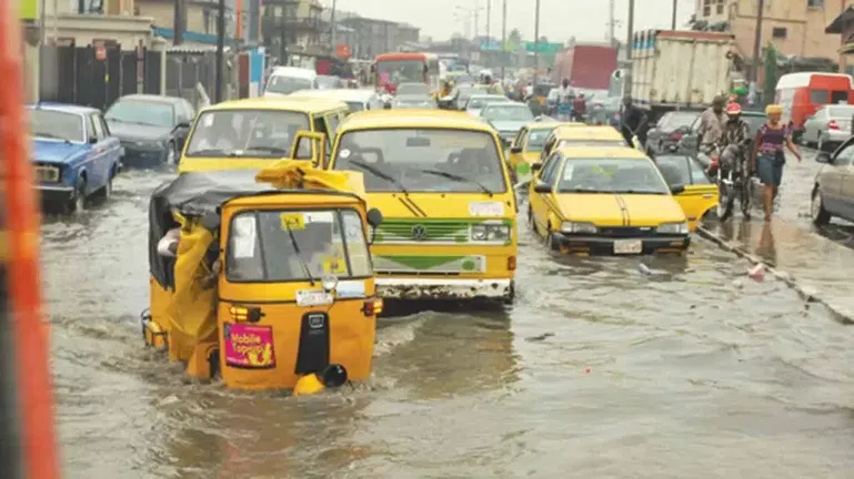Yellow commercial Danfo buses and tricycles (Keke Napep) struggling through deep floodwaters on a busy Ikorodu road in Lagos.