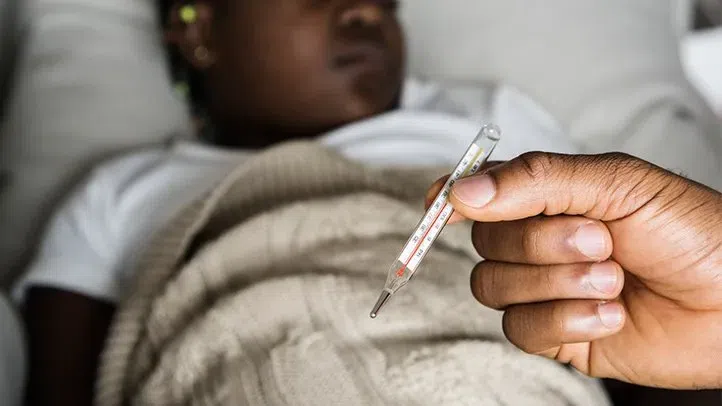 close-up of a person’s hand holding a glass clinical thermometer showing a high temperature, with a blurred child resting in the background under a blanket.