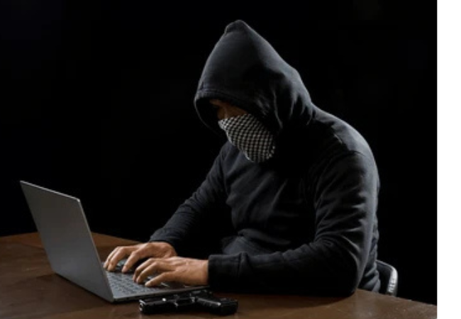 A cybercriminal wearing a black hoodie and face covering typing on a laptop with a handgun lying on a dark desk.