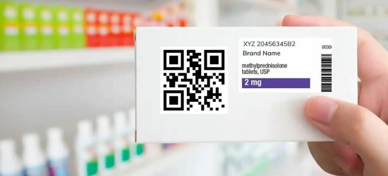 A person holding a white medicine box featuring a prominent scannable QR code on the packaging in a pharmacy.