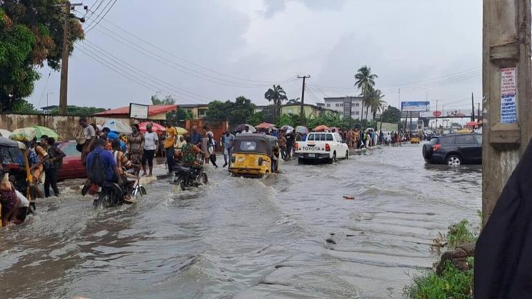 Commuters and keke napep tricycles are navigating a heavily flooded road in Agege, Lagos, during the rainy season.
