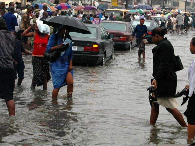 Lagos residents wading through deep flood waters on a busy road with submerged cars.