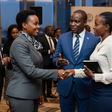 Three professionals networking at a business forum, with two women shaking hands while a man in a blue suit looks on and gestures during their conversation